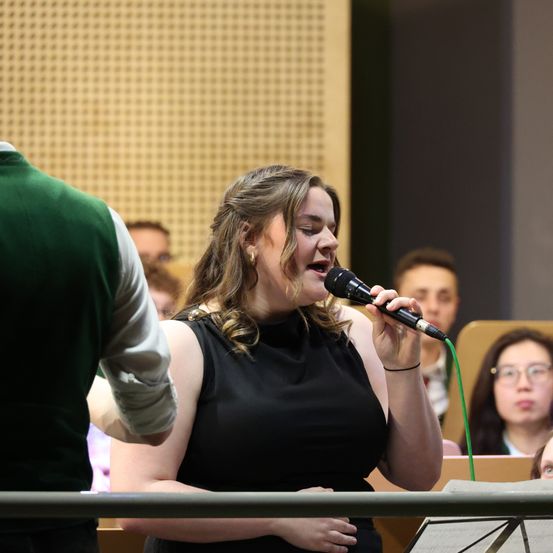 Eine Frau in einem schwarzen ärmellosen Oberteil singt in ein Mikrofon, während sie vor einer Gruppe von Menschen steht.