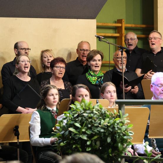 Ein Chor aus Erwachsenen und Kindern, in Schwarz gekleidet, singt in einem Raum mit grüner Wand und Holzstruktur. Sie halten Notenblätter und einige tragen Brillen.