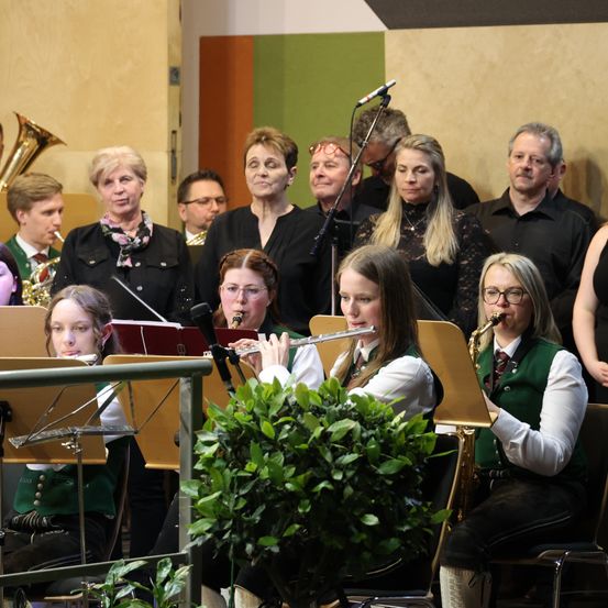 Eine Gruppe von Musikern, darunter eine Frau, die Flöte spielt, und eine andere, die Saxophon spielt, tritt in einem Raum auf, mit einer Pflanze im Vordergrund.