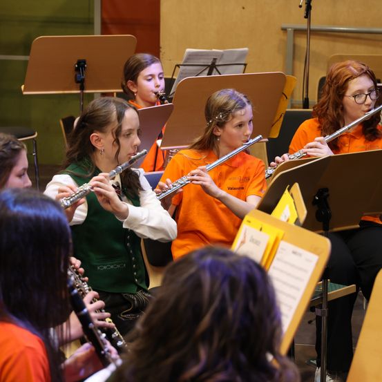 Eine Gruppe junger Mädchen in orangefarbenen Shirts spielt Flöten in einem Musikraum, wobei Notenständer und Notenblätter sichtbar sind.