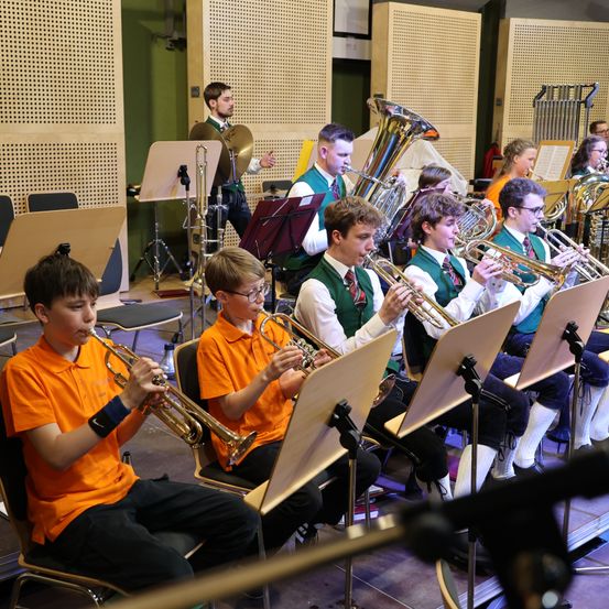 Eine Gruppe junger Musiker in orangefarbenen Hemden spielt Trompeten in einem Musikraum. Dahinter spielen andere Posaunen und Tubas.