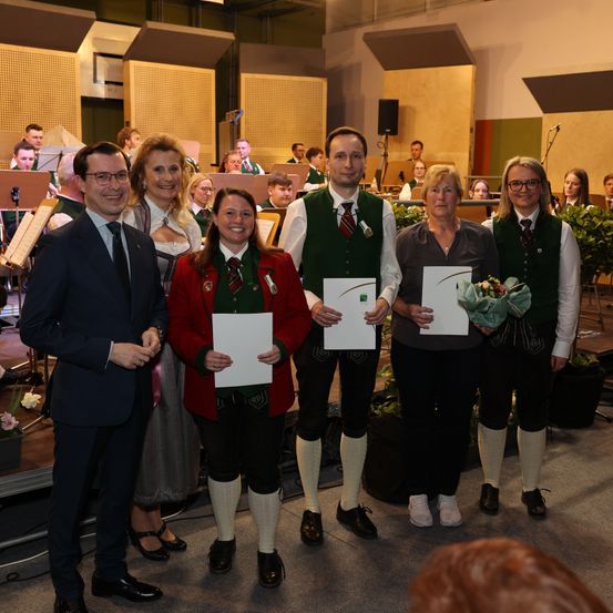 Eine Gruppe von Menschen in traditioneller Kleidung steht zusammen und hält Urkunden und Blumen in einem Konzertsaal. Dahinter spielen Musiker Instrumente.