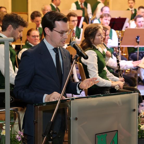 Ein Mann in Anzug und Krawatte spricht in ein Mikrofon an einem Rednerpult, mit einer Frau in Grün und Weiß hinter ihm und einer Band im Hintergrund.