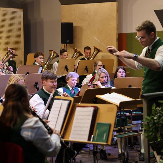 Ein Orchester spielt in einem Raum mit einem Dirigenten, verschiedene Instrumente wie Trompeten, Posaunen und Flöten sind sichtbar, und die Musiker tragen grüne Westen.