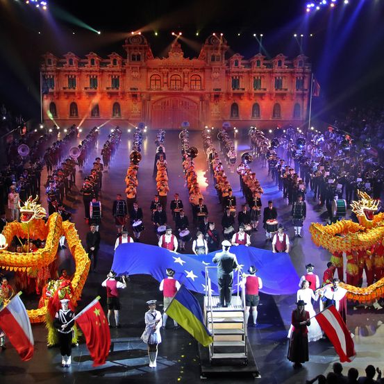 Ein großer Umzug auf der Bühne mit einem großen Gebäude im Hintergrund. Viele Menschen in Uniformen marschieren mit Flaggen und Musikinstrumenten. Eine große blaue Flagge mit dem Logo der Europäischen Union steht in der Mitte.