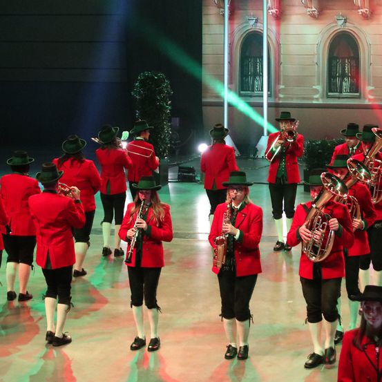 Eine Gruppe von Musikern in roten Uniformen und grünen Hüten spielt auf der Bühne mit verschiedenen Musikinstrumenten. Die Bühne ist gut beleuchtet mit grünen und roten Lichtern.