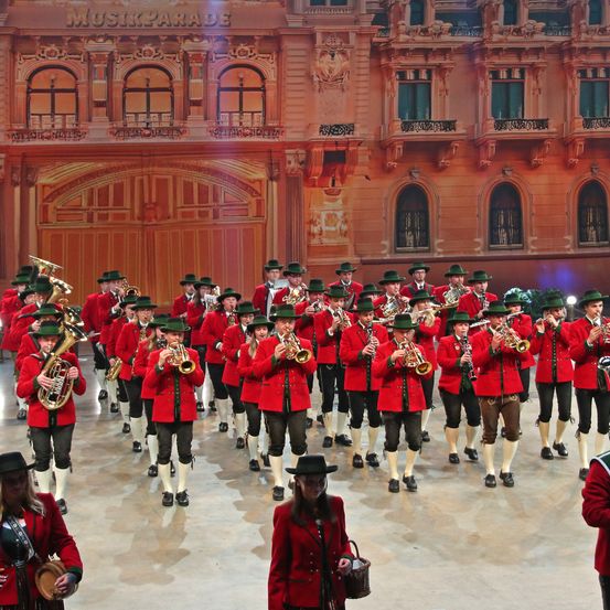 Ein Blasorchester in roten Uniformen spielt vor einem großen Gebäude mit kunstvoller Architektur. Die Musiker spielen verschiedene Instrumente, darunter Trompeten und Tuben.