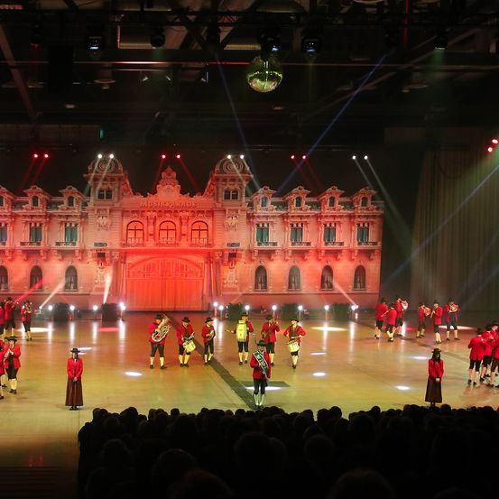 Eine Gruppe von Künstlern in roten Uniformen spielt Musikinstrumente auf einer Bühne mit rotem Hintergrund und einem großen Gebäude im Hintergrund.