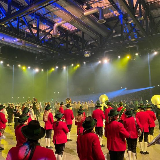 Eine große Gruppe von Menschen in roten Uniformen und grünen Hüten tritt mit Musikinstrumenten auf der Bühne unter farbigen Bühnenlichtern auf.