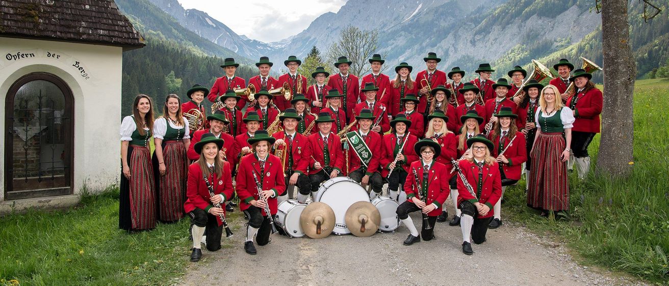 Eine Musikkapelle in traditionellen roten und grünen Uniformen posiert für ein Foto vor einem Berghintergrund. Einige sitzen mit Instrumenten, während andere dahinter stehen.