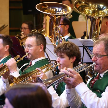 Eine Gruppe von Musikern in grünen Westen und weißen Hemden spielt Blechblasinstrumente in einer Aufführungsumgebung, wobei der Fokus auf Trompeten und Tuben liegt.