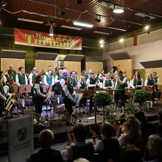 Eine Blaskapelle spielt auf der Bühne, mit Musikern in grünen und weißen Uniformen, umgeben von Topfpflanzen und einem Banner mit der Aufschrift 'Musikverein Turnau'. Das Publikum klatscht und schaut zu.