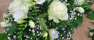 Ein weißes Blumenarrangement mit Rosen, Babys Atmung und grünen Blättern steht auf einer Holzoberfläche. Links ist ein goldener Gießkanister.