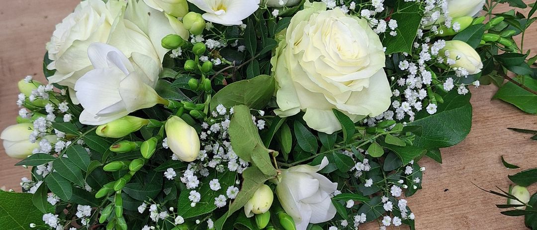 Ein weißes Blumenarrangement mit Rosen, Babys Atmung und grünen Blättern steht auf einer Holzoberfläche. Links ist ein goldener Gießkanister.