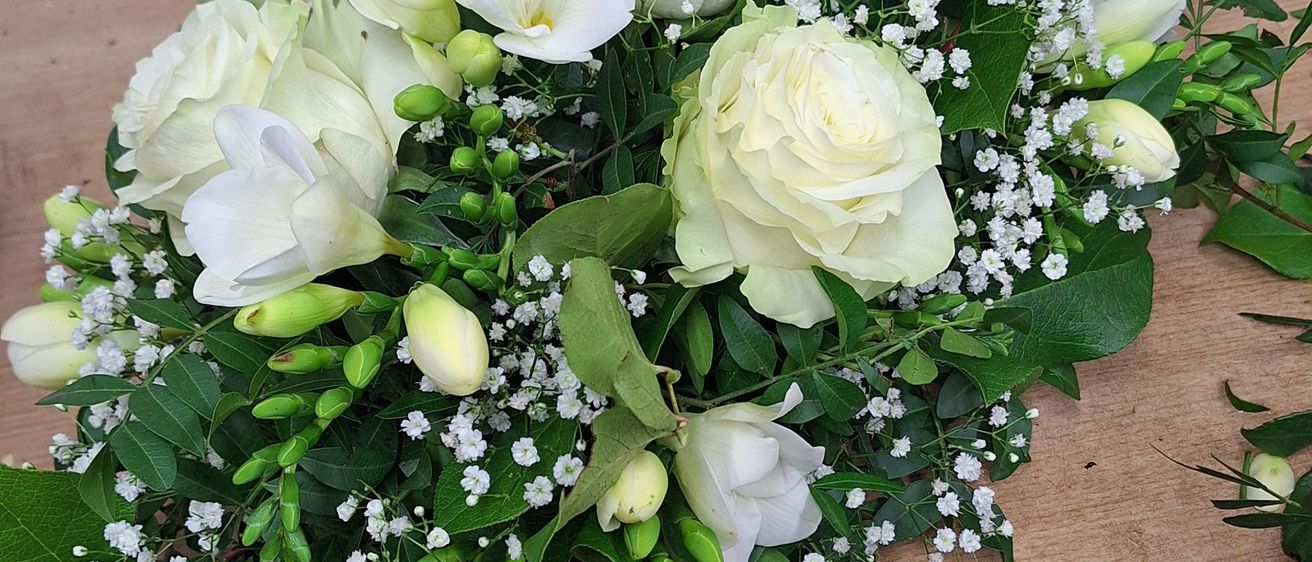 Ein weißes Blumenarrangement mit Rosen, Babys Atmung und grünen Blättern steht auf einer Holzoberfläche. Links ist ein goldener Gießkanister.