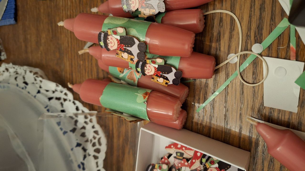 Eine Reihe roter Kerzen mit grüner Umwicklung und Comic-Charakter-Designs darauf steht auf einer Holzoberfläche. Darunter enthält ein Kasten mehr farbenfrohe, pilzförmige Gegenstände.