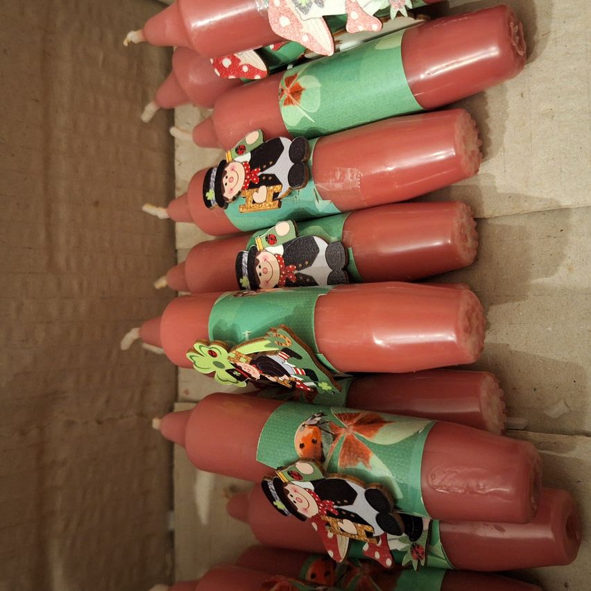 Eine Reihe roter Kerzen mit grünen und weißen Verpackungen ist in einer Schachtel angeordnet. Jede Kerze hat festliche Illustrationen wie einen Schneemann, eine Marienkäfer und eine Kuh.