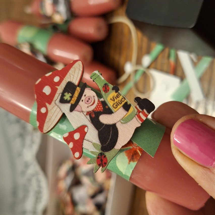 Eine Nahaufnahme eines rosa Objekts mit einem Holz-Schweinchen-Aufkleber, der einen Pilzhut, eine rote Schleife und Text auf Deutsch trägt. Das Objekt wird von einer Hand mit pinkem Nagellack gehalten.