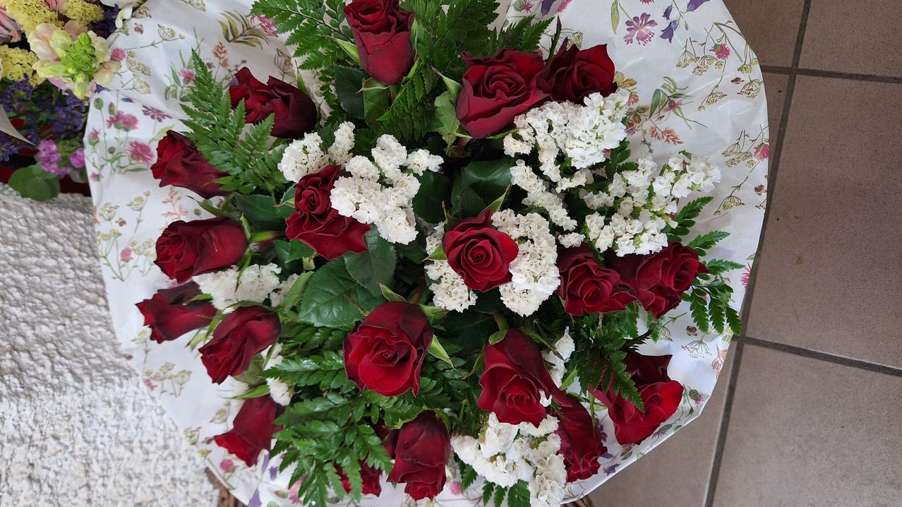 A beautiful bouquet of deep red roses and white flowers is arranged on a floral tablecloth. The roses are complemented by lush green leaves.