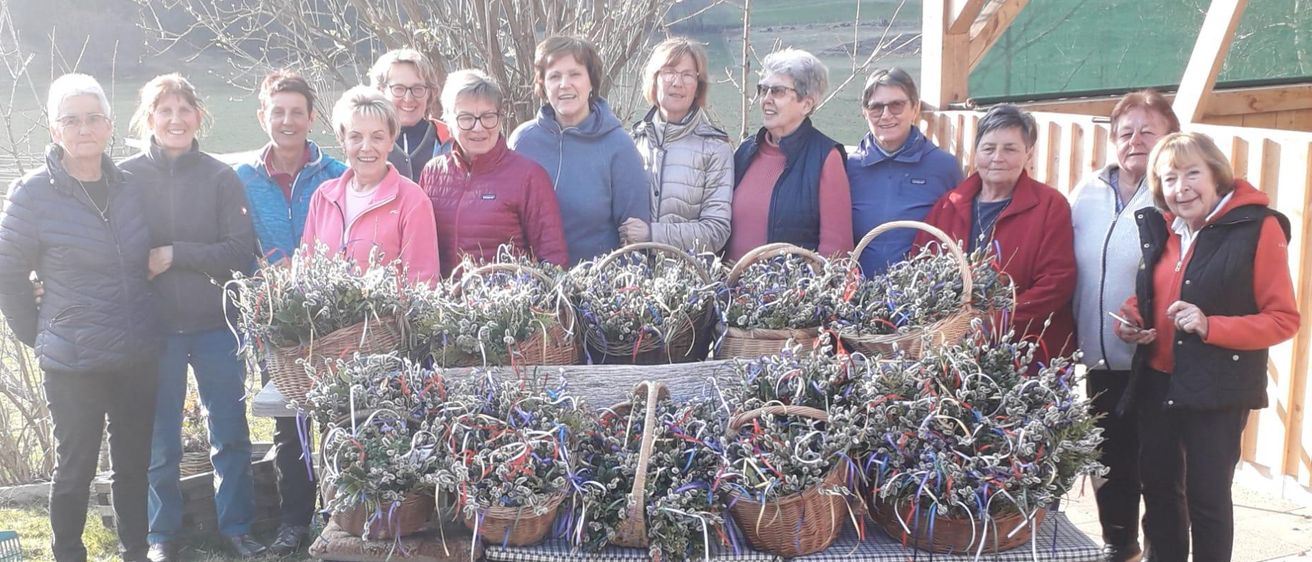 Eine Gruppe von Frauen steht hinter einem Tisch mit Blumenkörben und lächelt für ein Foto. Der Tisch ist mit einer karierten Tischdecke bedeckt.