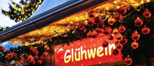 Ein Weihnachtsstand mit einem roten Schild, das Glühwein steht, umgeben von roten Kugeln und beleuchtet von gelben Lichtern, mit einem Weihnachtsbaum im Hintergrund.