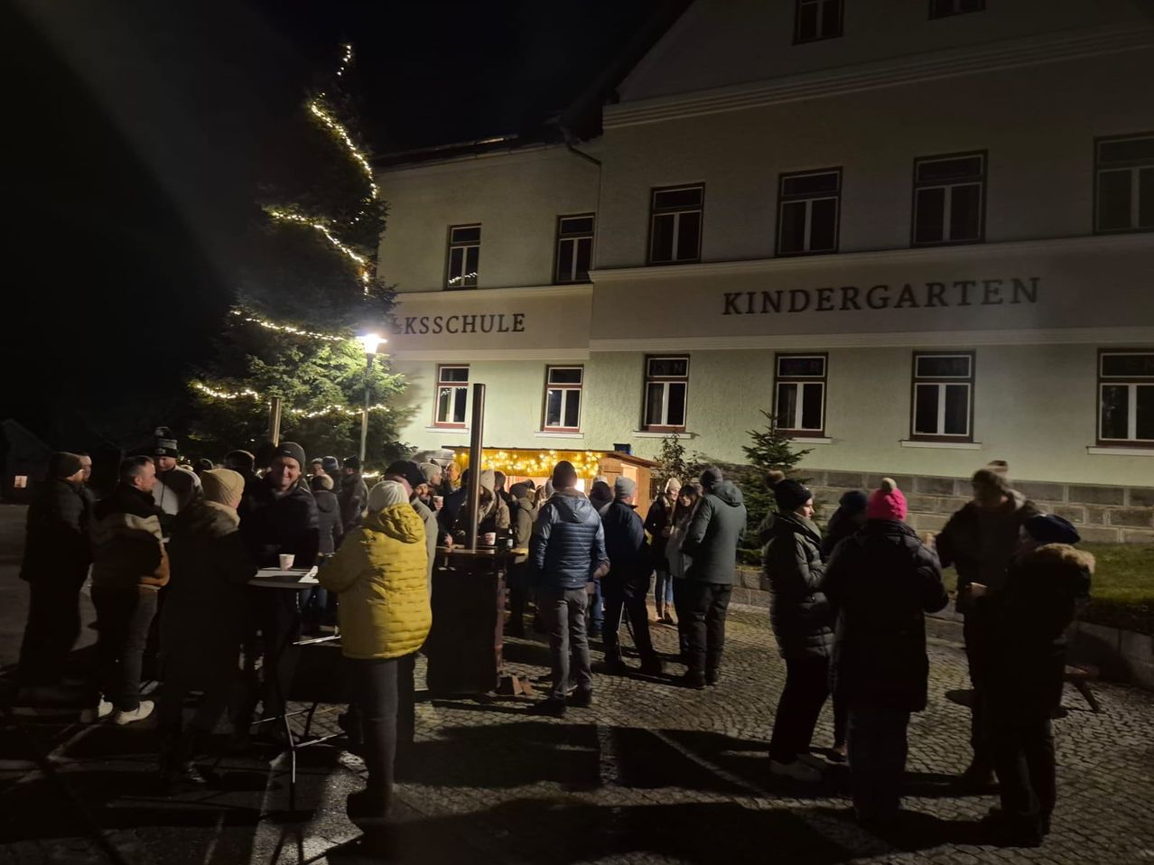 Eine Menschenmenge versammelt sich bei Nacht vor einem Gebäude mit der Aufschrift Kindergarten. Einige tragen Winterkleidung und Mützen. Ein beleuchteter Weihnachtsbaum steht vor dem Gebäude.