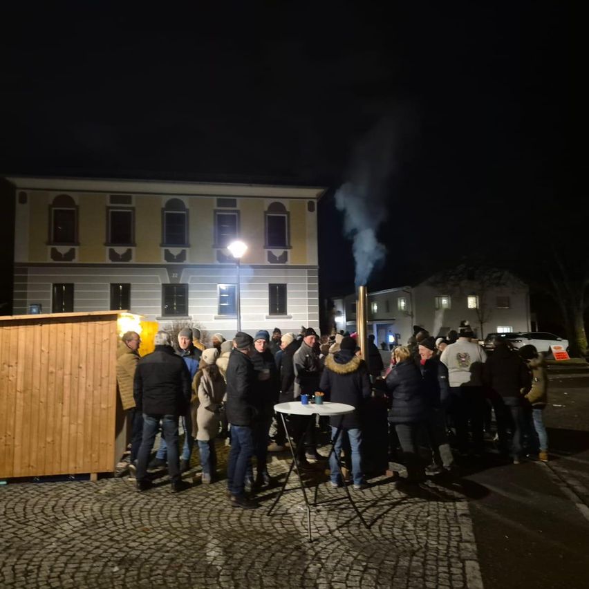 In der Nacht versammeln sich eine Gruppe von Menschen auf einem Platz, möglicherweise für eine Gemeinschaftsveranstaltung. Eine Holzhütte und ein Tisch mit Tassen sind vorhanden. Rauch steigt von einem Pfosten in der Mitte auf.