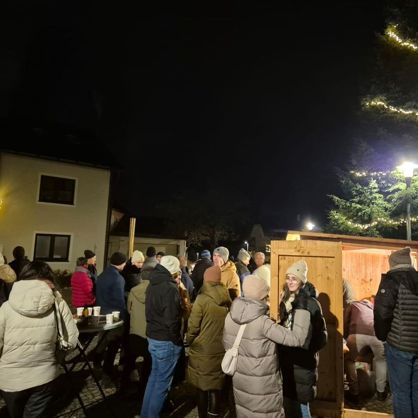 Bei Nacht versammeln sich Menschen um eine mit Lichtern geschmückte Holzstruktur, vielleicht für eine Feierlichkeit. Einige tragen Mützen und Mäntel, und in der Nähe steht ein Tisch mit Tassen. Ein Weihnachtsbaum und eine Straßenlaterne verleihen dem Fest eine weihnachtliche Atmosphäre.