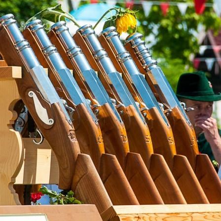Eine Reihe von Holz- und Silberschusswaffen, die auf einem Holzständer im Freien ausgestellt sind, mit einem Mann in einem grünen Hut, der dahinter steht.