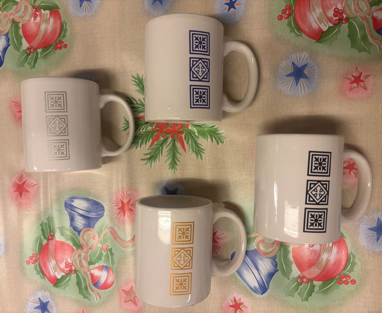 Fünf weiße Kaffeebecher mit verschiedenen Designs sind auf einer festlich dekorierten Tischdecke ausgestellt. Die Designs umfassen geometrische Muster, Blumenmotive und abstrakte Formen. Die Becher sind in einer Reihe angeordnet.