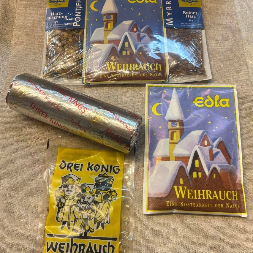 Auf einem Tisch sind verschiedene Weihnachtsweihrauchprodukte zu sehen. Eines ist eine silberummantelte Zylinder, der mit 'Drei Könige' beschriftet ist. Daneben befindet sich ein gelbes Päckchen mit einer Illustration von drei Königen. Zwei blau beschriftete Tüten zeigen 'Weihrauch' mit einer Hausillustration.