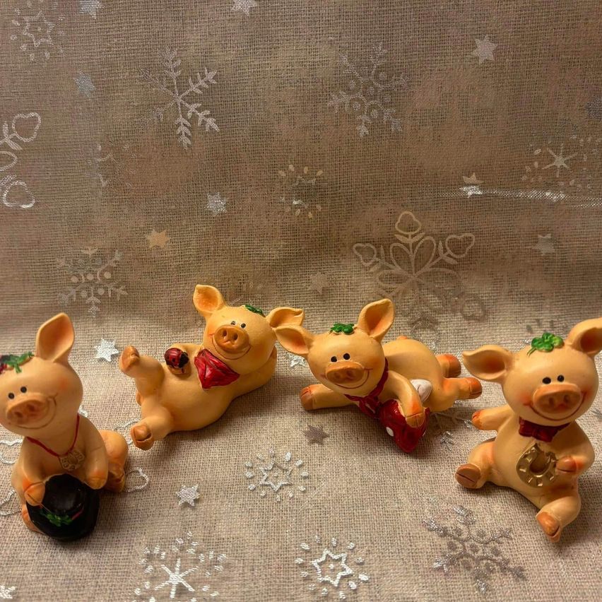 Fünf kleine Schweinefiguren sind auf einem weihnachtsstern-gemusterten Stoff angeordnet. Jedes Schwein trägt eine festliche Schleife und hält ein kleines Objekt. Sie sind in einem Halbkreis angeordnet, wobei ein Schwein eine rote Schleife und ein anderes eine Goldmünze trägt.