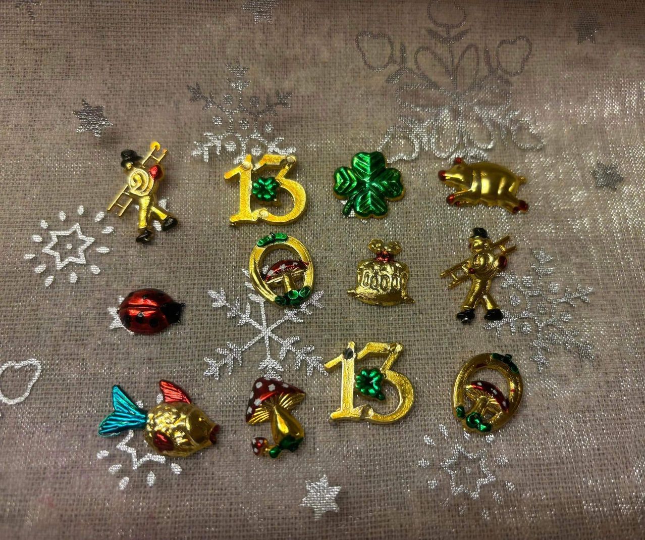 Eine Sammlung goldener Broschen mit den Zahlen 13, 15 und 16, die Symbole wie Glocke, Schwein, Pilz, Kleeblatt und Fisch zeigen. Die Broschen liegen auf einem grauen Tuch mit weißen Schneeflocken.