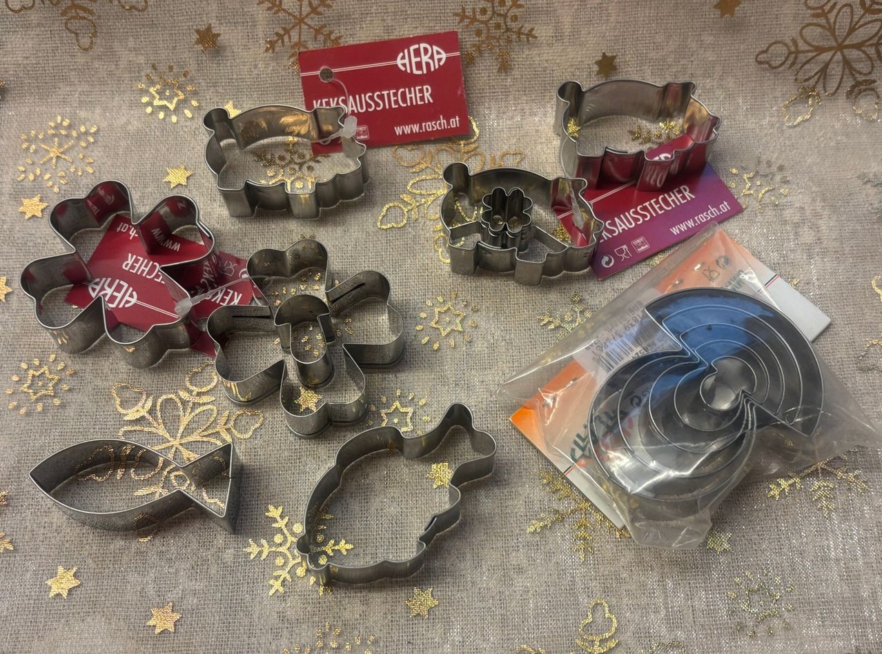 Verschiedene rostfreie Stahlplätzchenformen mit unterschiedlichen Formen sind auf einem Jutehintergrund mit goldenen Schneeflocken ausgestellt. Ein rotes Schild mit dem Wort 'HERD' ist vorhanden.