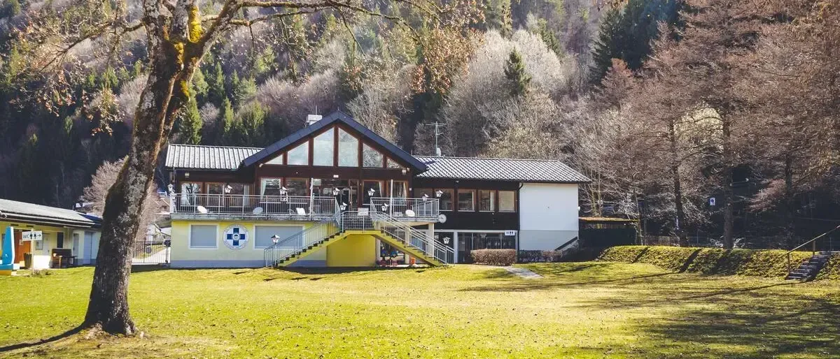 Ein Gebäude mit einem blau-weißen Emblem steht auf einer Rasenfläche, mit einem Baum und einer Treppe, die zu ihm führt. Berge sind im Hintergrund sichtbar.