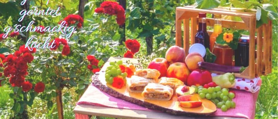 Ein Picknicktisch mit verschiedenen Früchten, Gebäck und einer Flasche Wein in einem Garten mit roten Blumen und einem Holzkasten. Das Bild hat einen Wasserzeichen mit dem Text 'Gaststand aus STRADEN'.