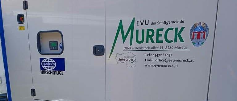 Ein weißer Anhänger mit einem Generator, der Mureck beschriftet ist, ist geparkt. Der Generator hat Logos und Kontaktdaten, einschließlich Telefonnummer und E-Mail.