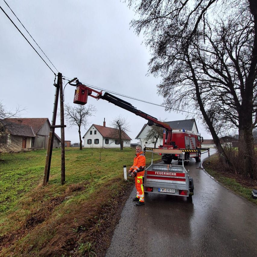 Ein Mann in Sicherheitsausrüstung steht neben einem Kran, der einen Strommast repariert. Der Kran ist auf einer nassen Straße geparkt, mit Häusern und Bäumen im Hintergrund.