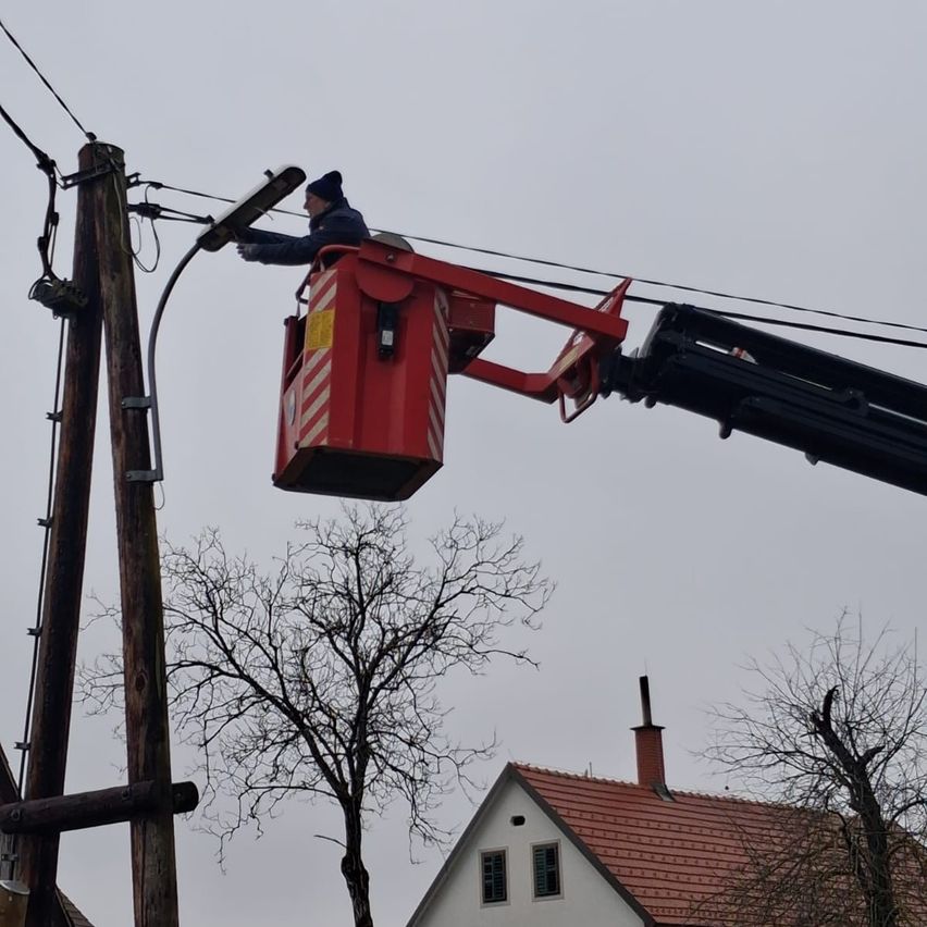 Ein Arbeiter in einem Arbeitskorb justiert elektrische Drähte über einem Haus mit einem roten Dach. Das Haus ist von kahlen Bäumen umgeben.