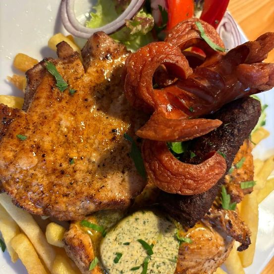 Ein Teller mit gegrilltem Hähnchen, Pommes und verschiedenen Gemüsesorten. Das Hähnchen hat eine knusprige Außenseite und ist mit Kräutern garniert. Es gibt eine cremige Dip-Sauce und eine Garnitur aus Zwiebeln.