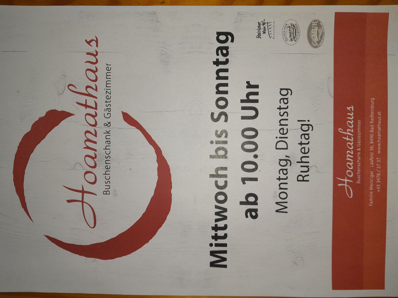 Ein weißes Holzschild mit rotem Text, das 'familienhaus' und 'Bäder und Gästezimmer' besagt. Darunter sind die Öffnungszeiten 'Montag, Dienstag, Mittwoch, Donnerstag, Freitag, Samstag, Sonntag von 10:00 bis 18:00 Uhr'. Auf der rechten Seite befinden sich auch zwei Logos.