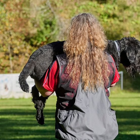 A woman carries a black dog on her back in a grassy area with trees in the background.
