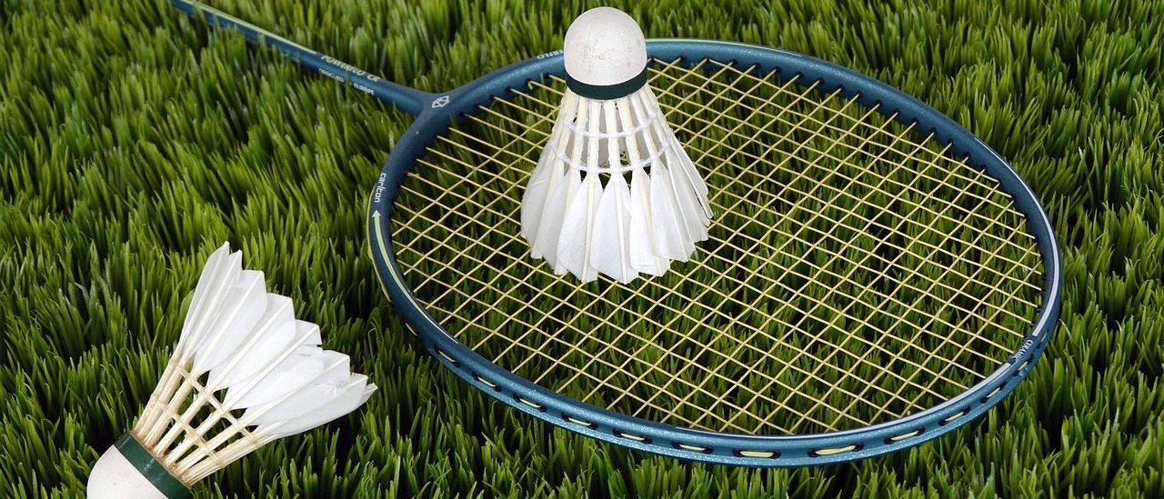 Ein blauer Badmintonschläger mit einem weißen Federball darauf liegt auf grünem Gras, mit einem weiteren weißen Federball in der Nähe.