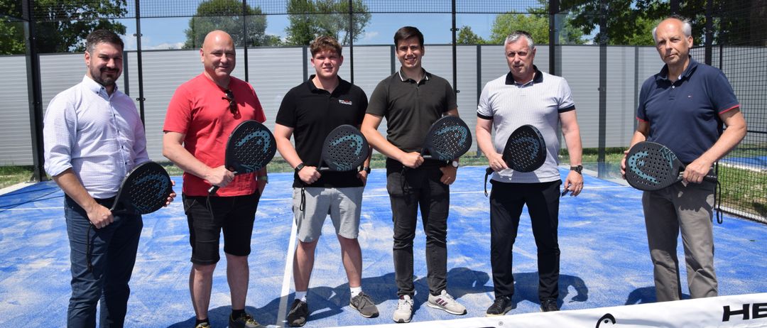 Fünf Männer auf einem blauen Tennisplatz, Tennisschläger in der Hand, lächeln und posieren für ein Foto. Der Platz hat ein Netz und ein weißes Banner mit dem Wort HEAD. Dahinter steht ein Zaun.