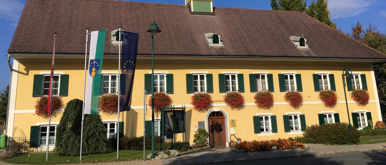 Ein gelbes Gebäude mit einem braunen Dach und grünen Fensterläden hat einen Uhrenturm mit einem Kreuz oben. Flaggen und Pflanzen sind davor.