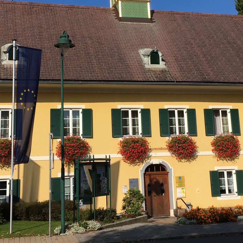 Ein gelbes Gebäude mit einem braunen Dach und grünen Fensterläden hat einen Uhrenturm mit einem Kreuz oben. Flaggen und Pflanzen sind davor.