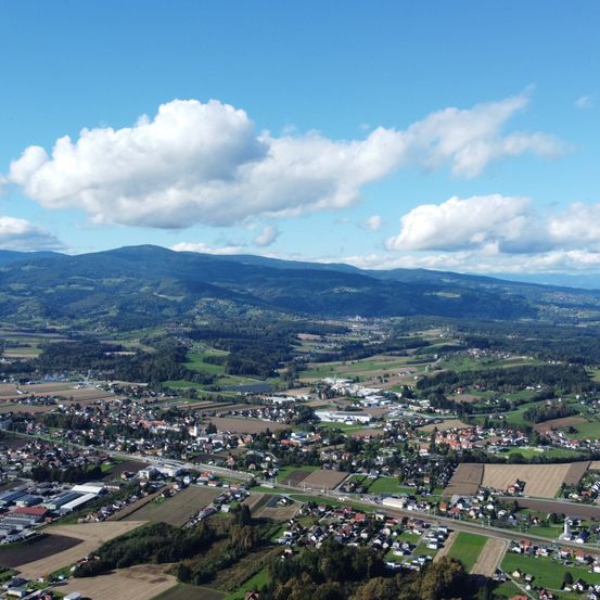 Luftaufnahme einer Stadt mit Gebäuden, Häusern und Straßen, umgeben von grünen Feldern, Bäumen und Bergen unter einem blauen Himmel mit Wolken.