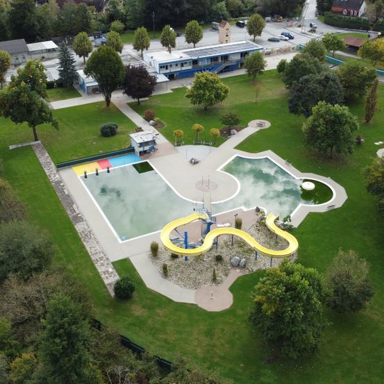 Eine Draufsicht eines Parks mit Schwimmbad, gelber Rutsche und verschiedenen Bäumen. Im Hintergrund sind Gebäude und Autos zu sehen.