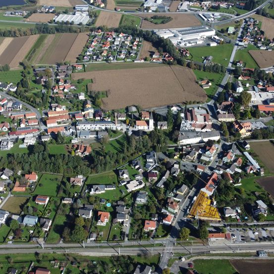 Luftaufnahme einer Stadt mit vielen Häusern, einige mit roten Dächern, umgeben von Feldern und Grün. Gebäude sind unter Bäumen verstreut. Eine große Fabrik ist in der Ferne sichtbar.