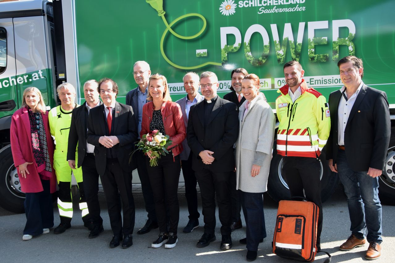 Eine Gruppe von Menschen in formeller Kleidung, einer hält Blumen, steht neben einem grünen Lastwagen. Ein Mann in einem gelben Sicherheitsjacke und einer orangefarbenen Tasche steht rechts.